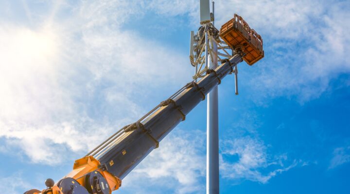 Crane with telescopic boom lift used as an aerial working platform. Worker install cellular base station with transmitters 3G, 4G, and antennas on cell tower on blue sky background.
